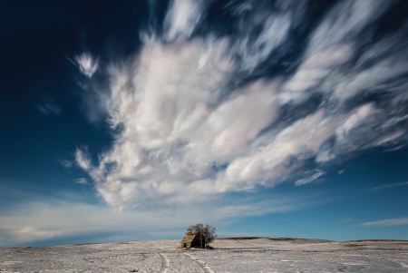 Small cabin in a snowy field with huge clouds above it.